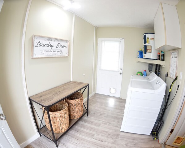 A laundry room with a wooden table, wicker baskets, a white washing machine, and a shelf with cleaning supplies.