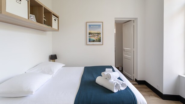 Habitació d'hotel amb un llit, un llençol blau, un quadre al mur i una prestatgeria de fusta sobre el llit.