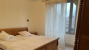 Una cama bien hecha con cabecera de madera, una mesita de noche y una ventana con cortinas traslúcidas.