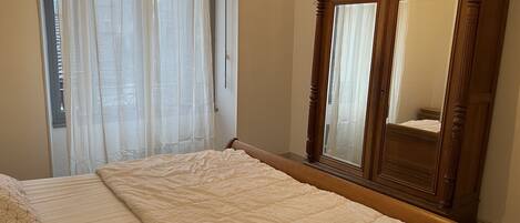 Un dormitorio con cama, armario de madera y ventana con cortinas traslúcidas.