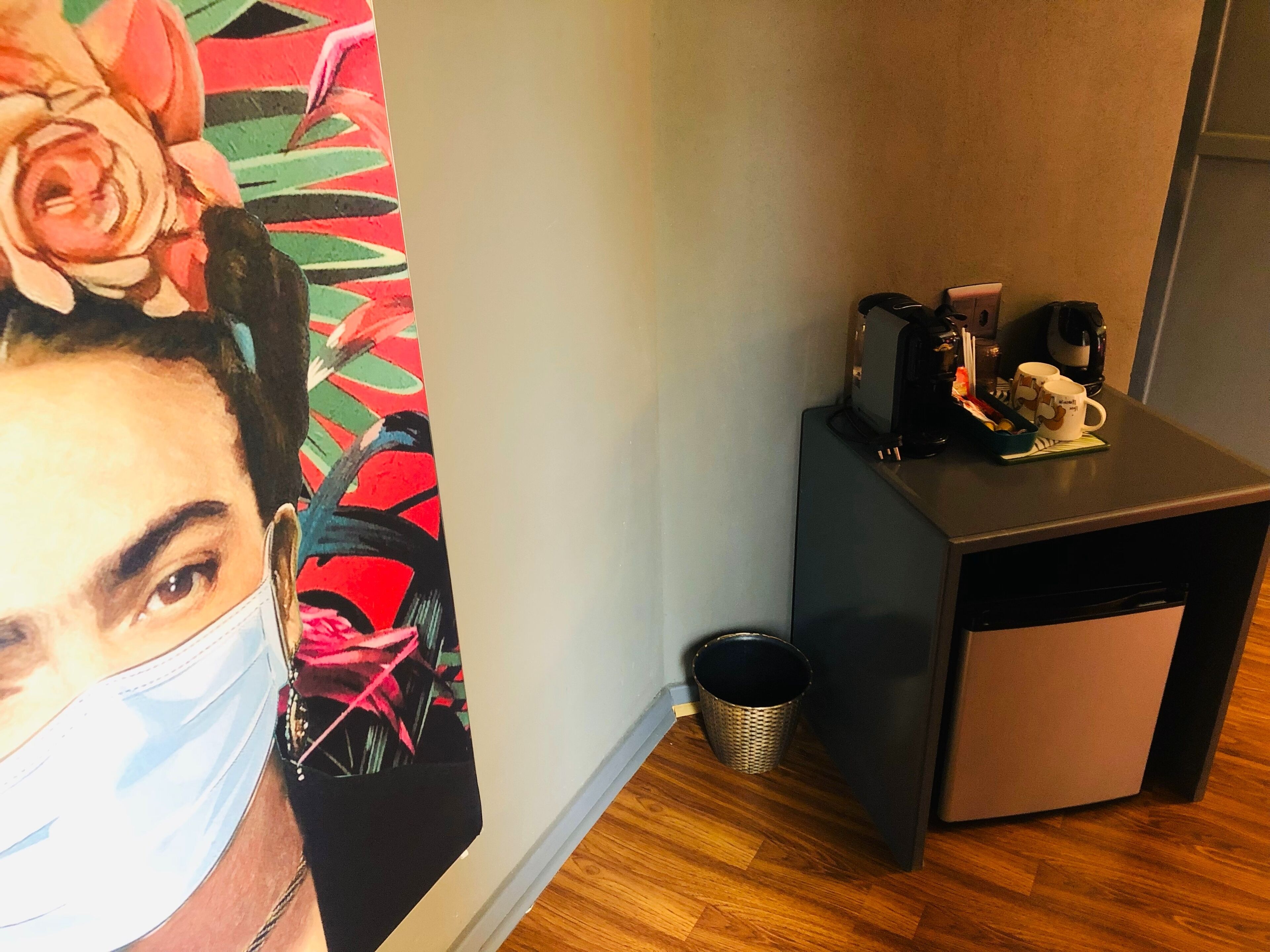 Une pièce avec un plancher en bois, un petit bureau avec une machine à café, une poubelle et un tableau coloré au mur.
