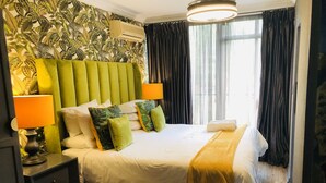 Hotellihuone, jossa on vihreäverhoiltu sängynpääty, valkoisilla lakanoiden peitetty sänky, vihreitä ja valkoisia tyynyjä, keltainen heittopeitto, yöpöytä lampun kanssa ja ikkuna, jossa on verhot.