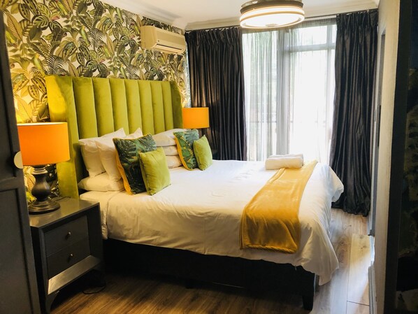 Habitación de hotel con cabecera tapizada en verde, cama con sábanas blancas, almohadas verdes y blancas, una manta amarilla, una mesita de noche con lámpara y una ventana con cortinas.
