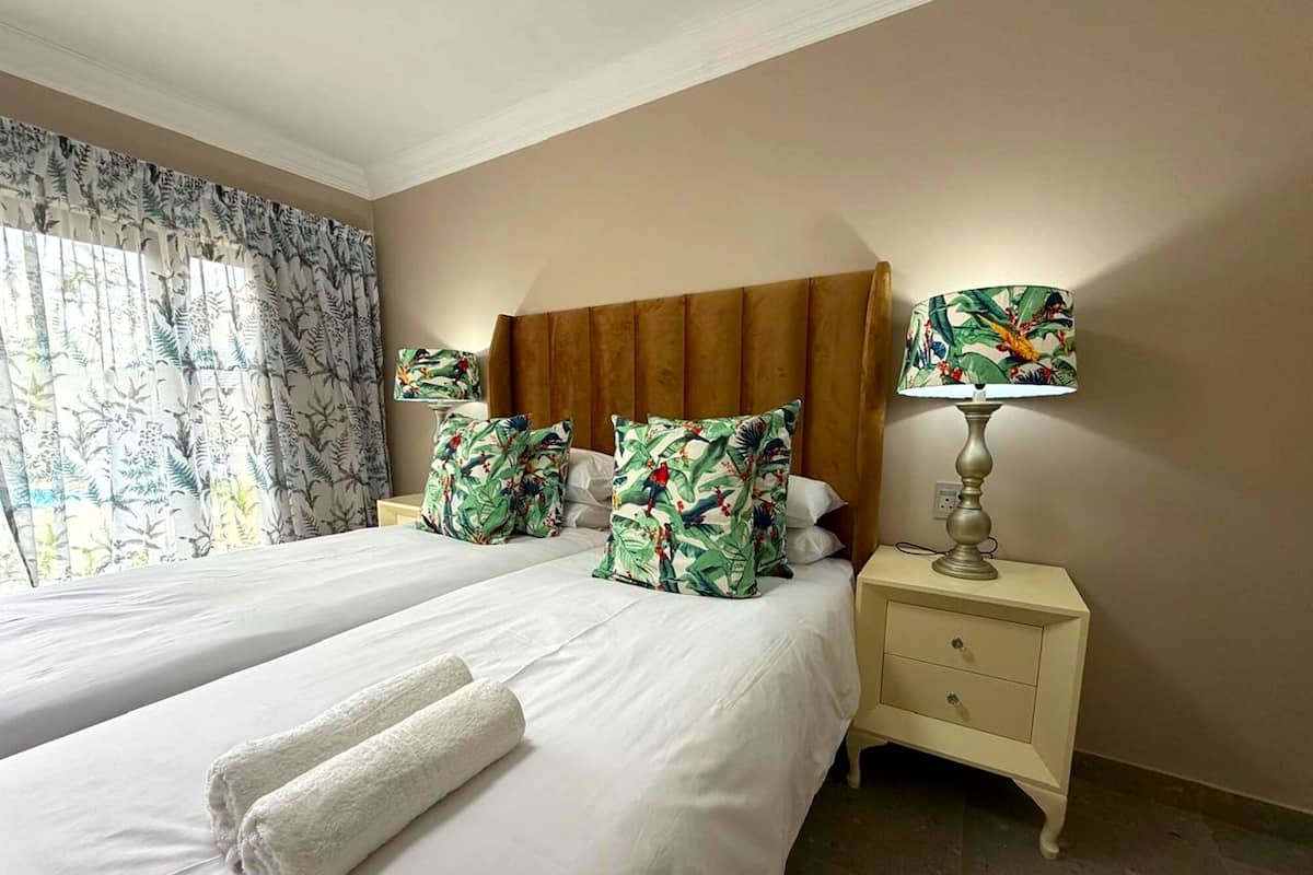 A hotel room with two beds, a nightstand with a lamp, and a window with curtains.