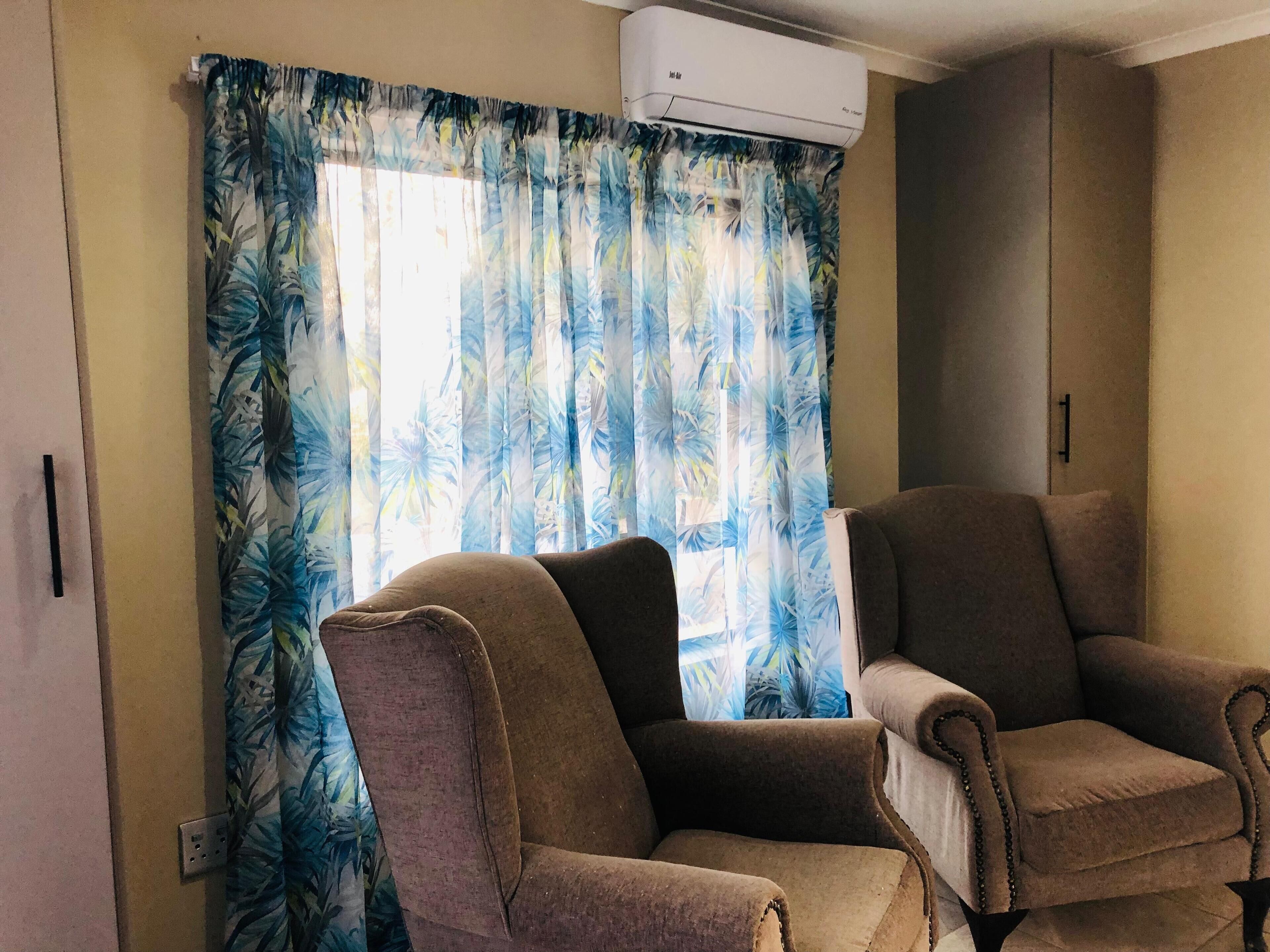 Una habitación con dos sillones marrones, una ventana con cortinas tropicales azules y un equipo de aire acondicionado en la parte superior.