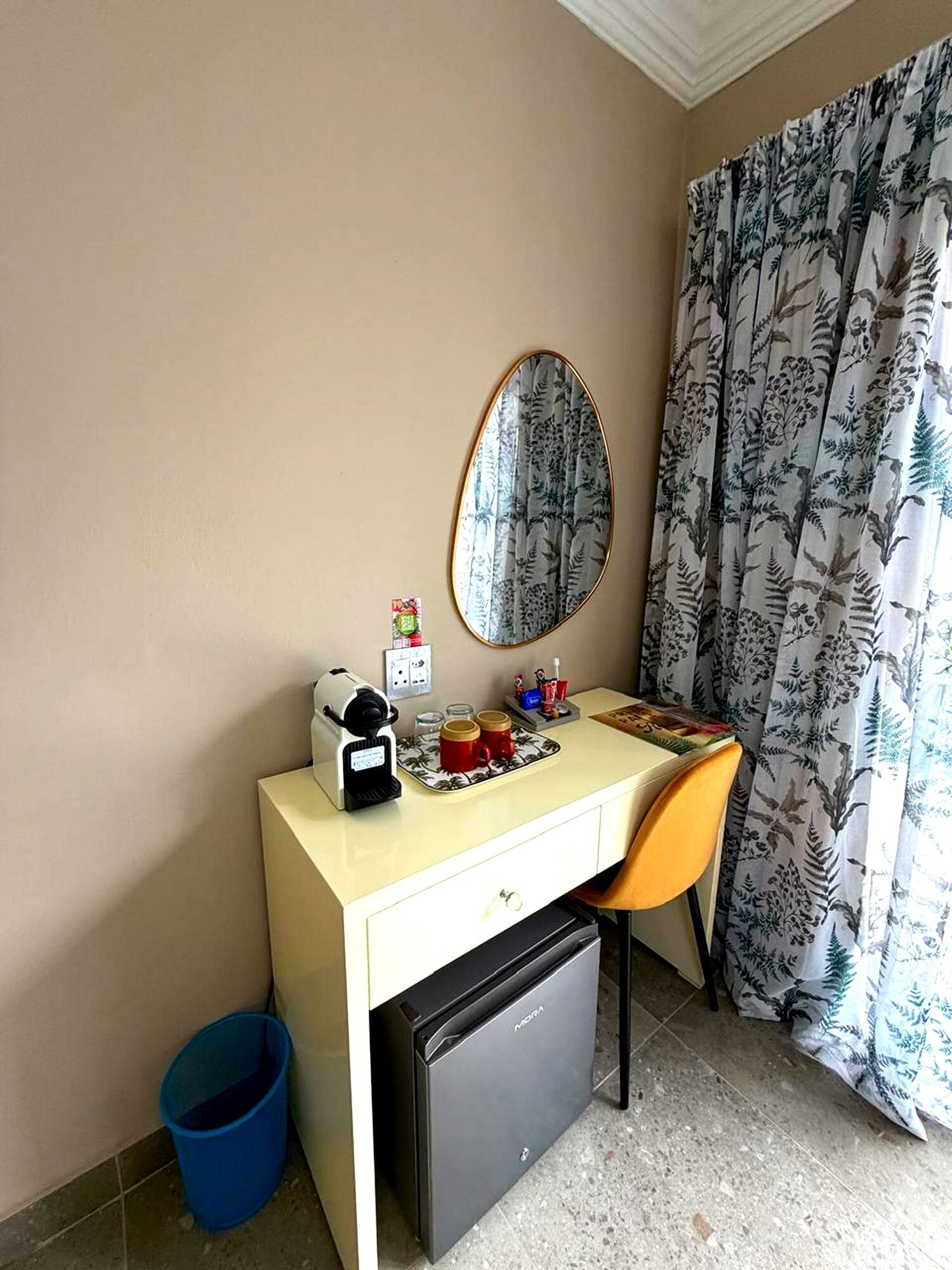 A compact hotel room with a small desk, a coffee maker, a mirror, and a compact refrigerator.