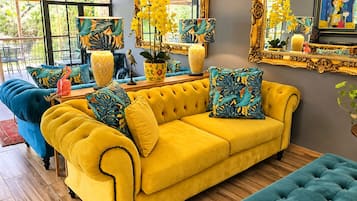 Ein Zimmer mit einem gelben Sofa, einem blauen Sessel und einem türkisfarbenen Hocker. Auf der Wand befinden sich dekorative Vasen und ein Spiegel.