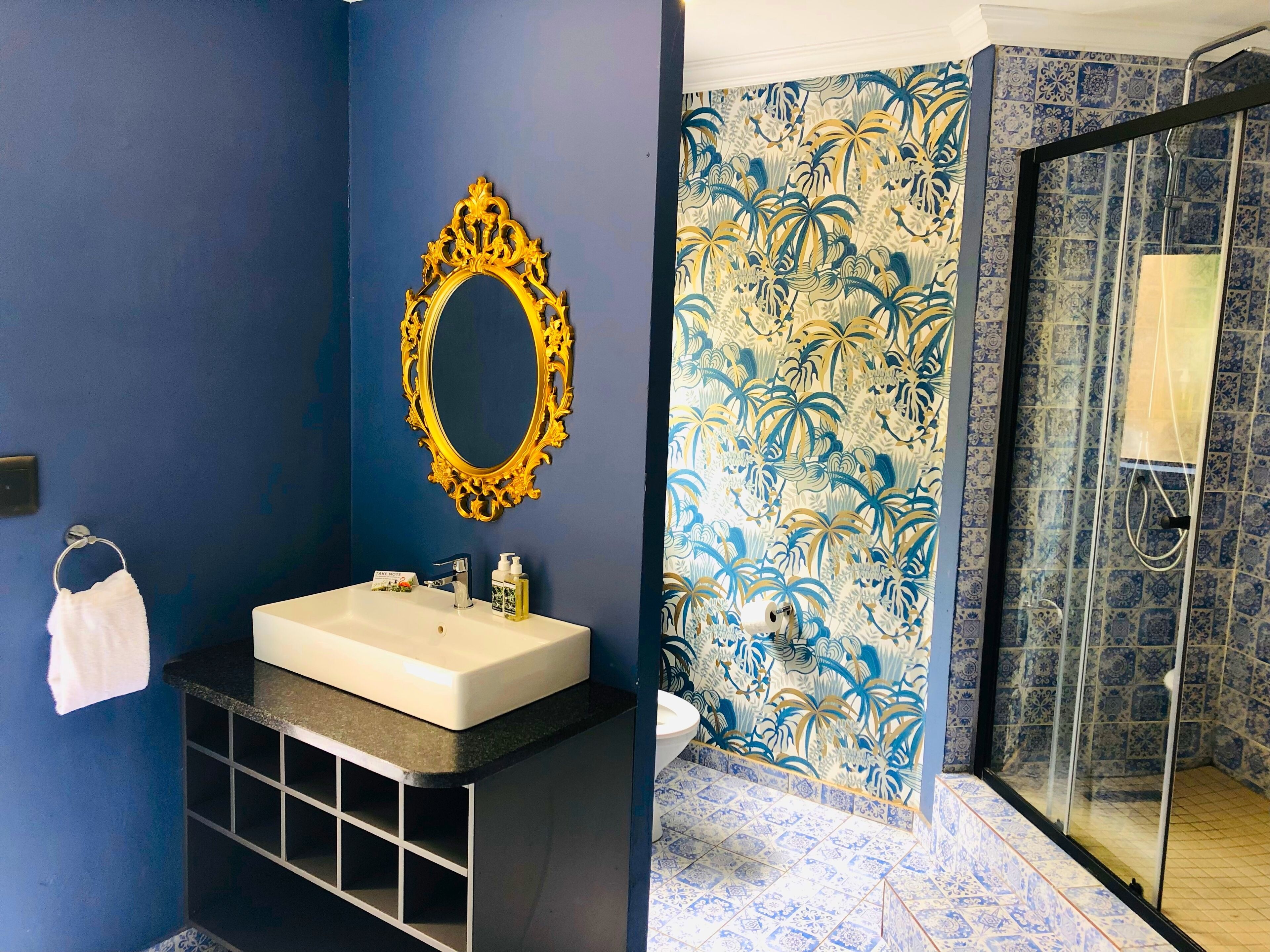 A bathroom with a white sink, a gold-framed mirror, a white towel, and a shower with blue patterned tiles.