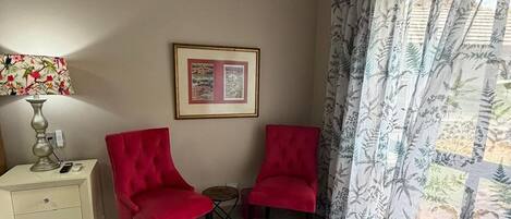 Ein Zimmer mit zwei roten Stühlen, einem kleinen Tisch, einem weißen Nachttisch mit Lampe, einem gerahmten Bild an der Wand und einem gemusterten Vorhang.