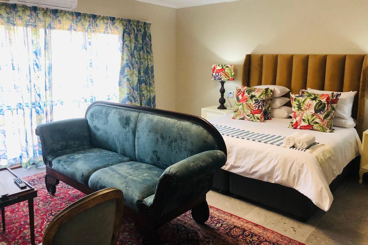 A hotel room with a blue sofa, a bed with floral pillows, a nightstand, and a lamp.
