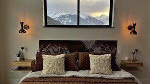 A bedroom with a large bed, two bedside tables, a window with a mountain view, and wall-mounted lamps.