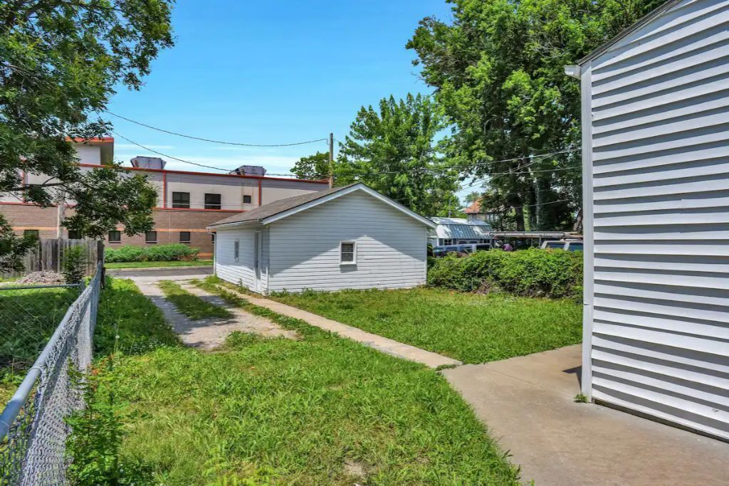 A small, single-story house with a pathway leading to the front door, surrounded by a fenced yard and adjacent to a larger building.