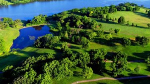 Oversigtsbillede af en golfbane med en sø, omgivet af frodig vegetation og træer.