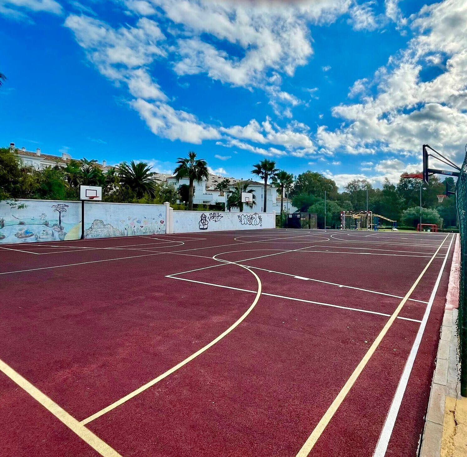 En utomhusbasketplan med rött underlag och vita linjer som markerar planen, omgiven av grönska och en klar himmel.