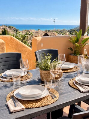 An outdoor dining setup with a view of the sea, featuring a table set with plates, glasses, and a bottle, surrounded by chairs and a potted plant.