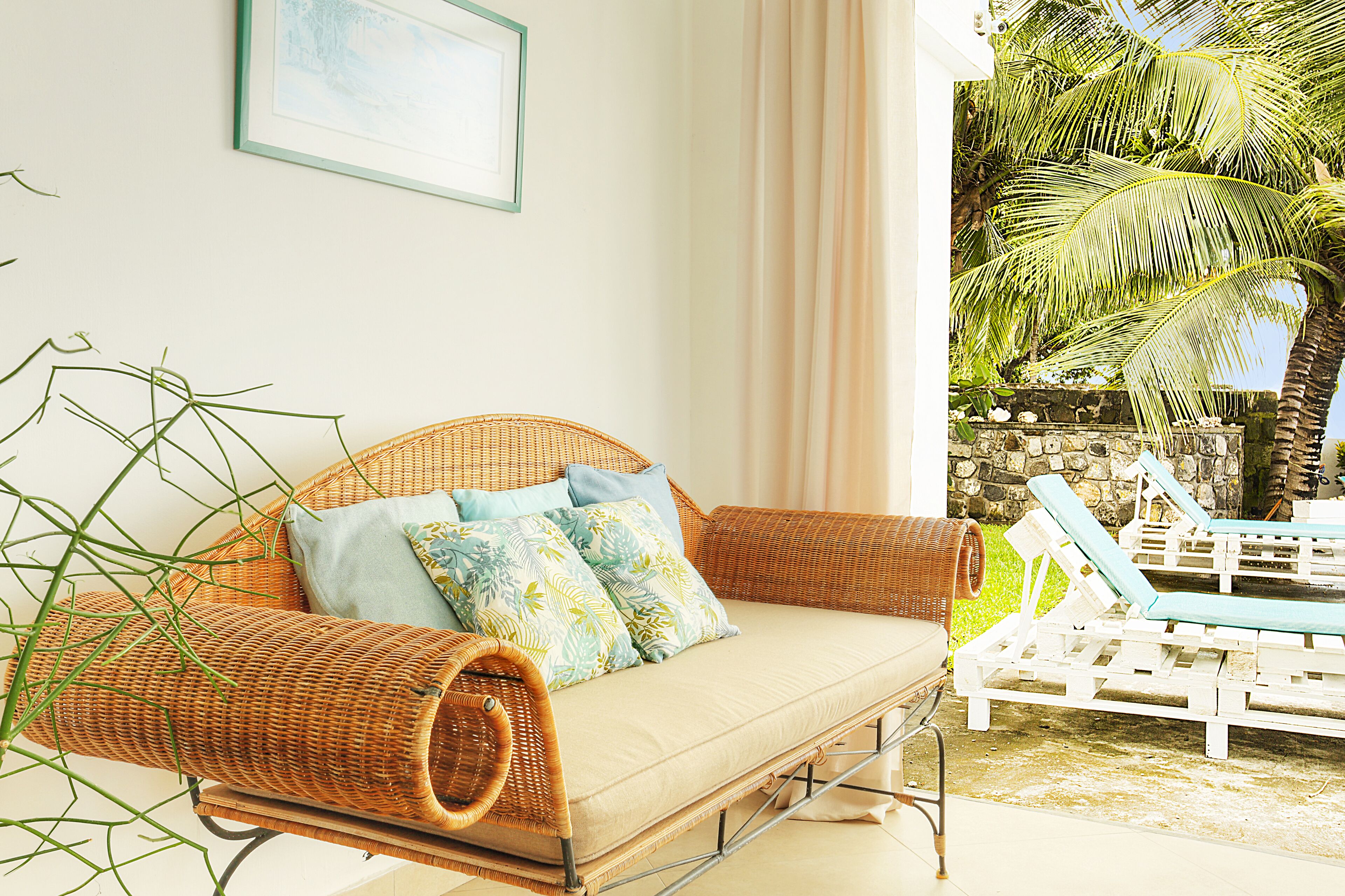 A wicker sofa with cushions, a glass table, and a view of a stone wall and palm trees.