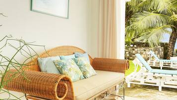 A wicker sofa with cushions, a glass table, and a view of a stone wall and palm trees.