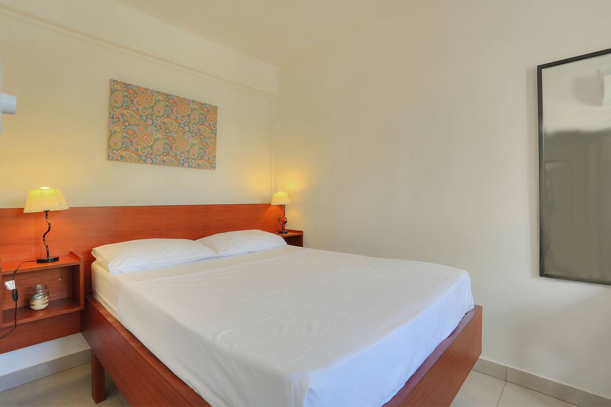 A neatly made bed in a hotel room with a wooden headboard, two bedside lamps, and a colorful abstract painting on the wall.