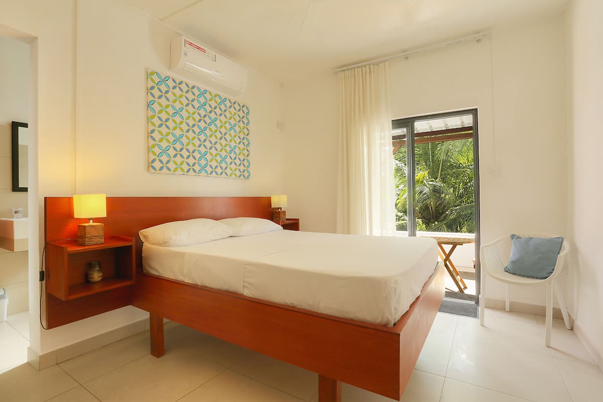 A hotel room with a wooden bed, a nightstand with a lamp, a patterned wall hanging, a window with curtains, and a view of greenery outside.