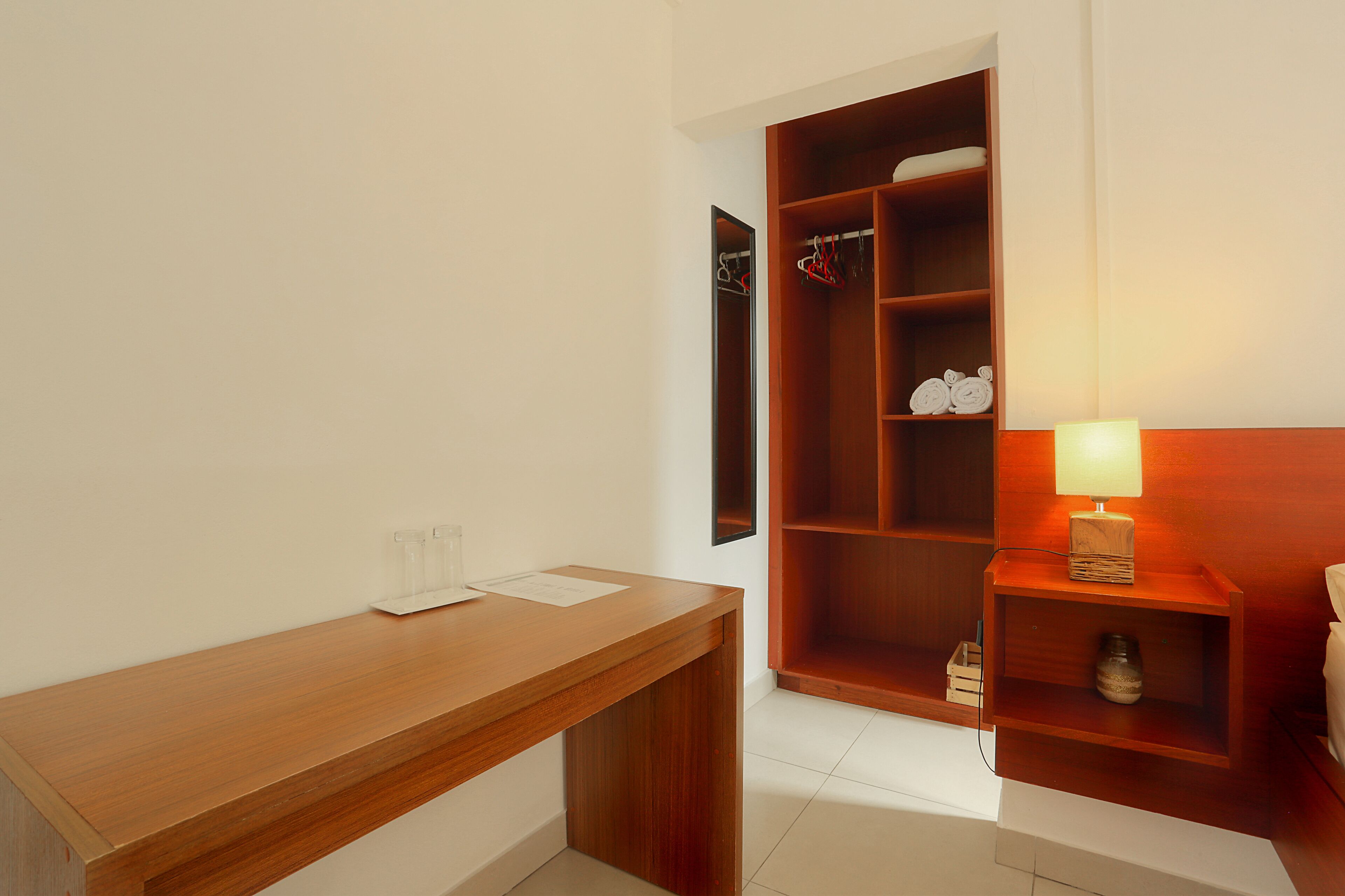 A hotel room with a wooden desk, a wardrobe with hanging clothes, and a bedside table with a lamp.