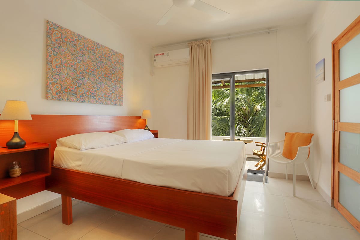 A hotel room with a wooden bed, two bedside tables with lamps, a large window with curtains, a chair, and a colorful abstract painting on the wall.