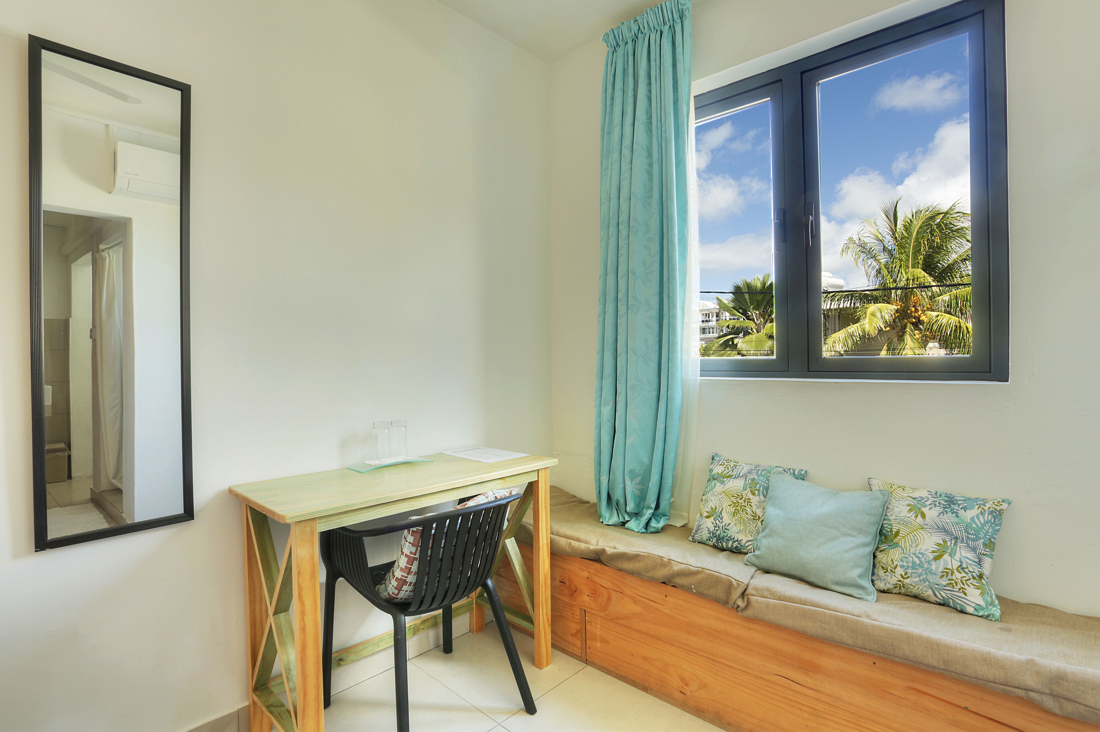 Una habitación con un escritorio de madera, una silla negra, una ventana con vistas a palmeras, un espejo y un banco con cojines estampados.