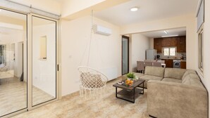 Un appartement moderne avec une porte vitrée donnant sur un balcon, un fauteuil suspendu, un canapé beige, une table basse avec des fruits, et une cuisine en arrière-plan.