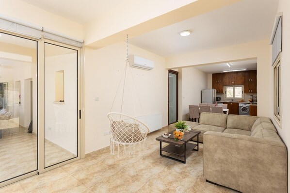 A modern apartment with a glass door leading to a balcony, a hanging chair, a beige sofa, a coffee table with fruit, and a kitchen in the background. - Villa Elite - Four Bedroom Villa, Sleeps 8 (Drouseia)