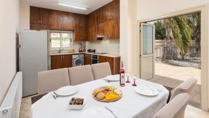 Une cuisine compacte avec des meubles en bois, un four encastré et un lave-linge. Une table à manger recouverte d’une nappe blanche est dressée avec des assiettes, des couverts et un plateau de fruits.