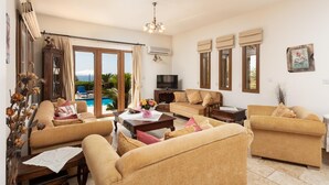 A spacious living room with a large sofa set, a coffee table, and a television. There is a view of a pool through the glass doors. - Villa Erato - Three Bedroom Villa, Sleeps 6 (Nea Dimmata)