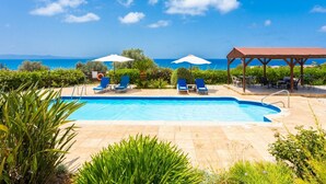 A pool with blue water, surrounded by lounge chairs and umbrellas, with a view of the sea and hills in the background. - Villa Erato - Three Bedroom Villa, Sleeps 6 (Nea Dimmata)