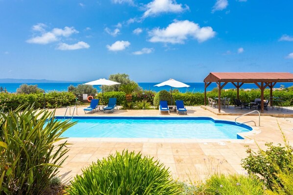 Une piscine à l’eau bleue, entourée de chaises longues et de parasols, avec une vue sur la mer et les collines en arrière-plan.