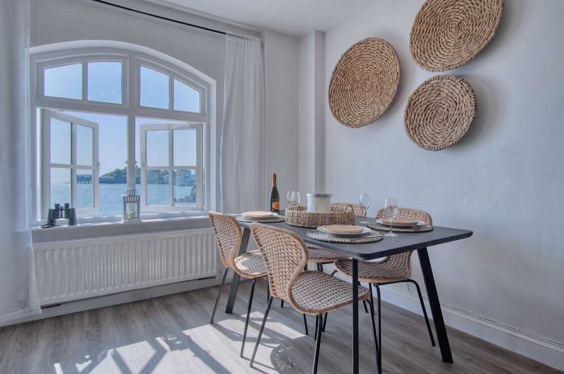 A dining area with a table set for four, wicker chairs, and a view of the sea through large windows.