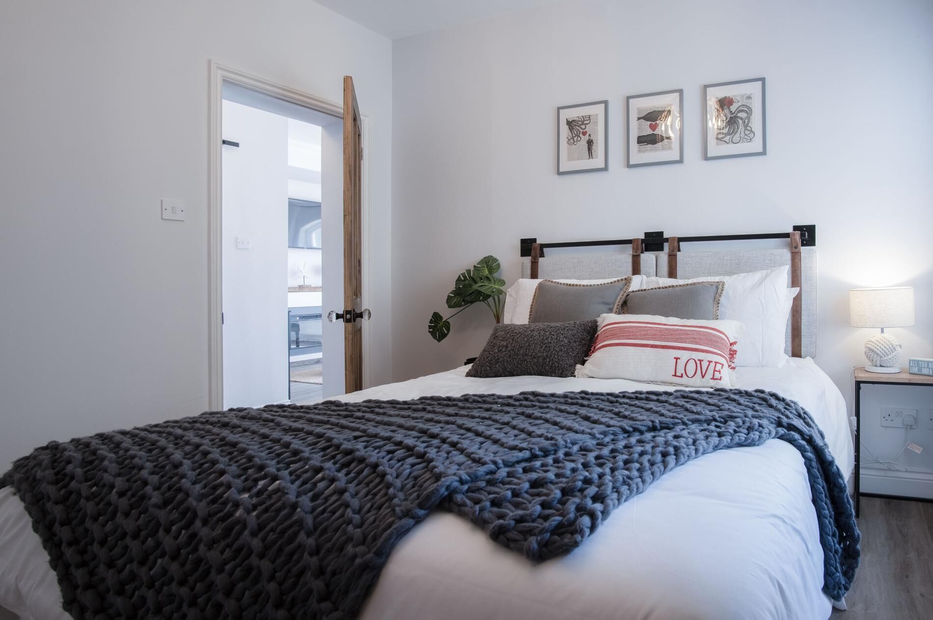 A neatly made bed with a dark knitted blanket, a pillow with the word "LOVE", and three framed artworks on the wall.
