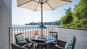 A balcony with wicker furniture, a table with a glass top, and a large umbrella. - The Sand Castle - 2 Bedroom Apartment - Tenby (Tenby)