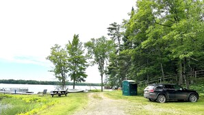 A gravel path leading to a campsite with picnic tables, a portable toilet, and a car parked nearby. - Lakefront 2BR Condo on Namakagon – Views, Trails, Pontoon Rentals & Fishing! (Cable)