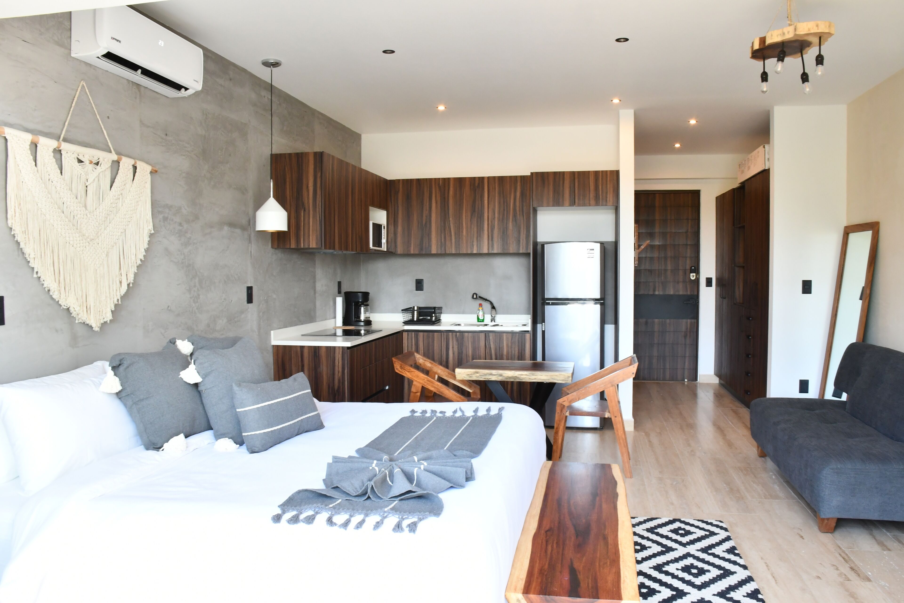 A modern hotel room with a bed, a wooden dining table, a kitchenette, and a gray sofa.