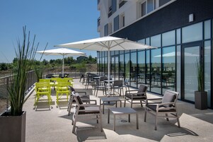 Un patio al aire libre con sillas amarillas y grises, sombrillas blancas y vistas a la ciudad a través de amplios ventanales.