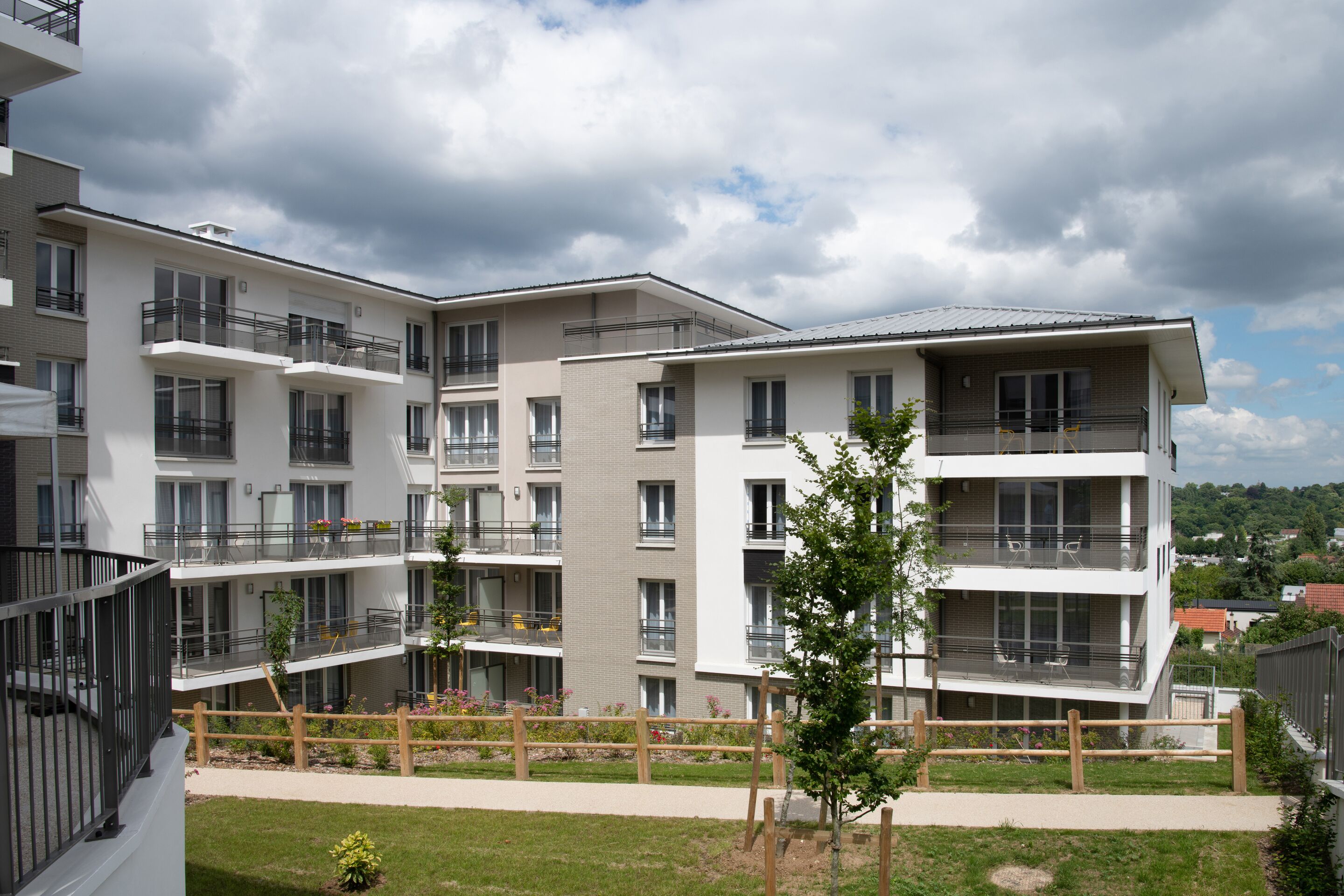 Un immeuble résidentiel à plusieurs étages, doté de balcons, d’une cour verdoyante et surplombant un ciel dégagé.
