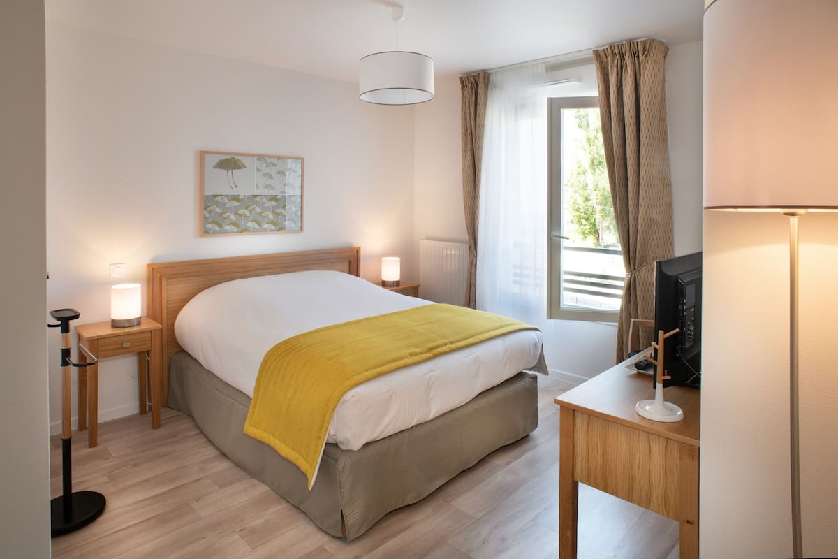 Quarto de hotel com cama, abajures, escrivaninha com televisão e janela com cortinas.