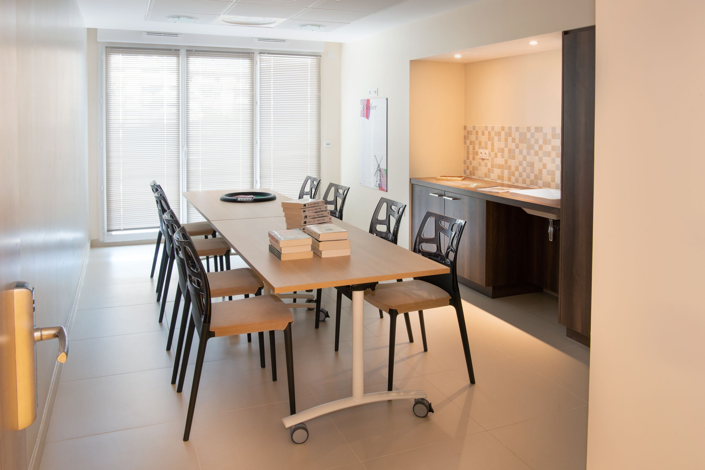 Uma sala de conferências moderna com uma mesa grande, cadeiras e uma área de cozinha com painel de azulejos.