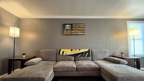 Uma sala de estar com sofá seccional, mesa de centro e duas mesas laterais com luminárias. Há uma obra de arte na parede acima do sofá.