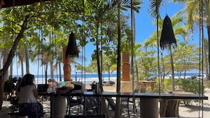 En udendørs siddeplads med borde og stole med udsigt over en strand med palmer og havet.