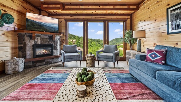 A cozy cabin interior with a stone fireplace, two blue armchairs, a blue sofa, and a large window offering a view of the outdoors. - Luxury Views, Sparkling Pool, Giant Theater Oh My! (Sevierville)
