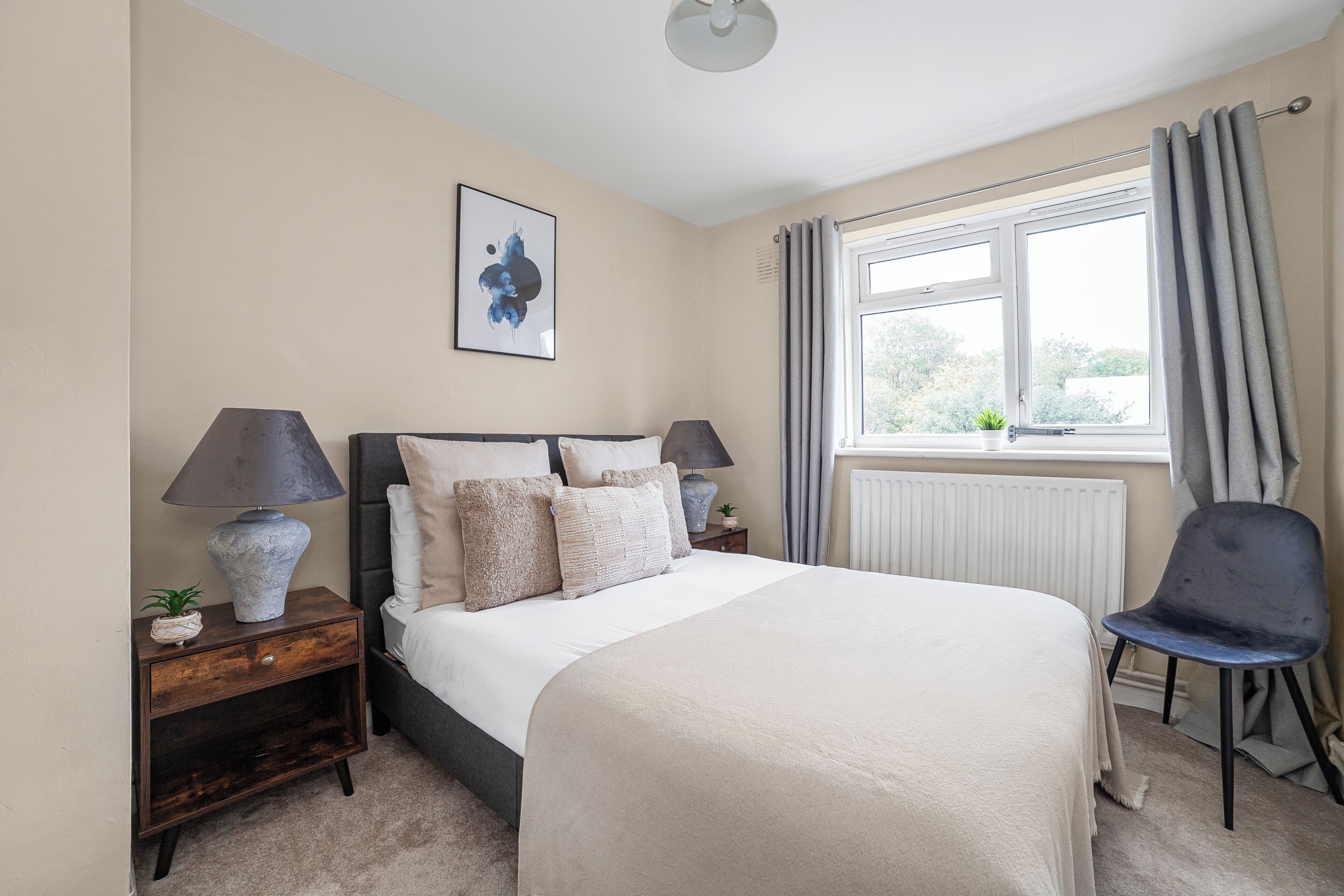 A bedroom with a large bed, two bedside tables, a chair, a window with curtains, and a radiator.