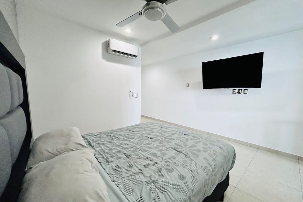 A bedroom with a bed, a ceiling fan, an air conditioner, and a mounted TV.
