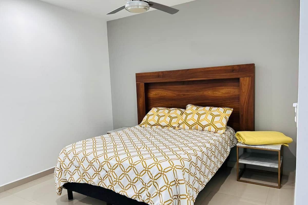 A bedroom with a bed, a wooden headboard, a bedside table with a yellow towel, and a ceiling fan.