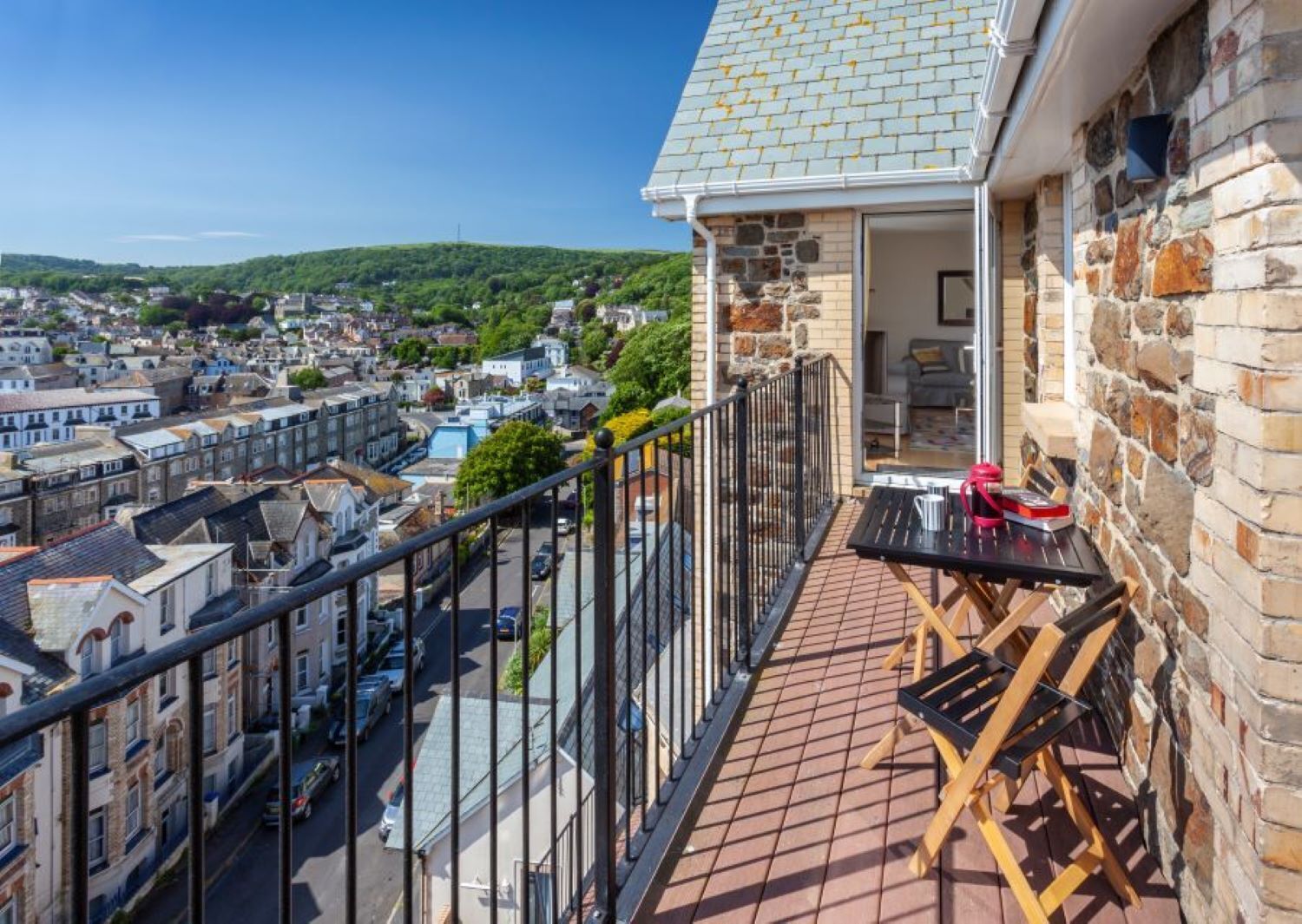 A balcony with a table and chairs, overlooking a residential area with houses and a hill in the background.