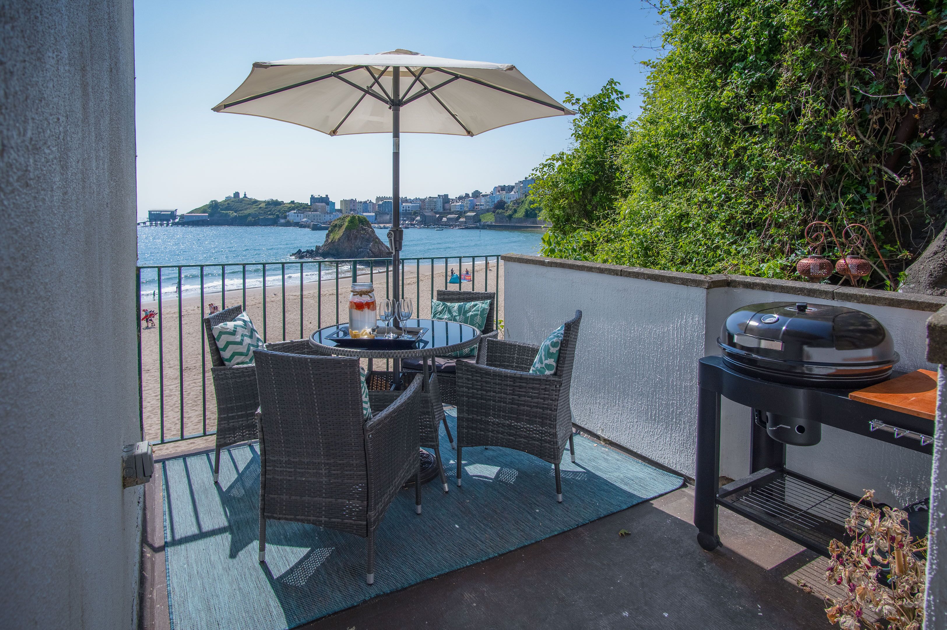 A balcony with wicker furniture, a grill, and a view of the beach and sea.