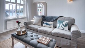 Un salon moderne avec un canapé gris, une table basse en bois et une vue sur une étendue d’eau grâce à de grandes fenêtres.
